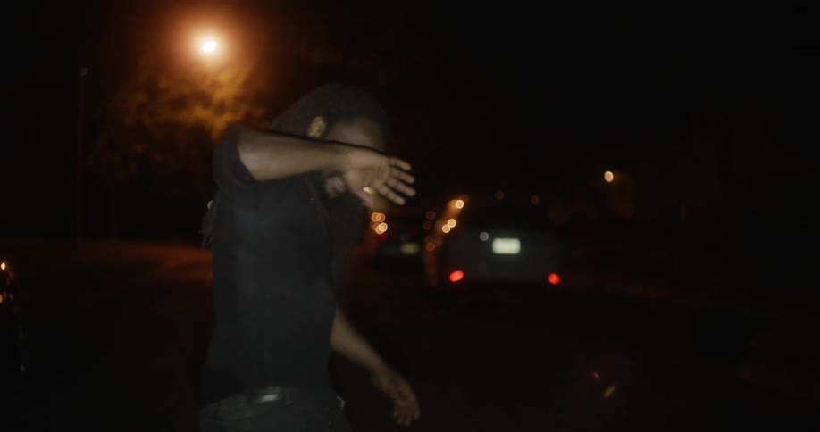 A man shades his eyes from a light as he walks and staggers on a street at night