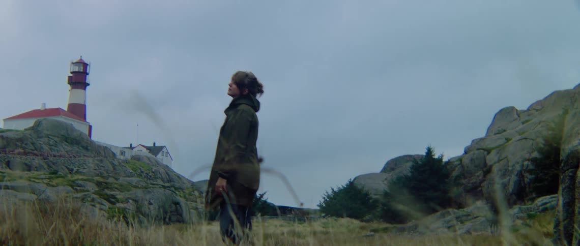 A woman stands in a field near a lighthouse and looks around