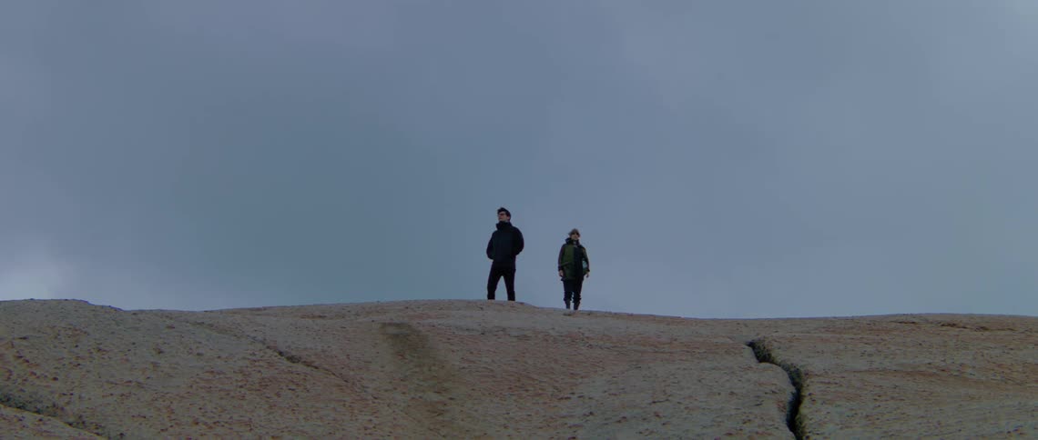 Two people wearing dark clothes are standing on top of a mountain hill