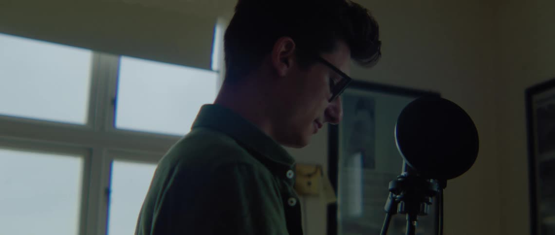 A man wearing glasses is talking to someone as he is looking down at something he is doing