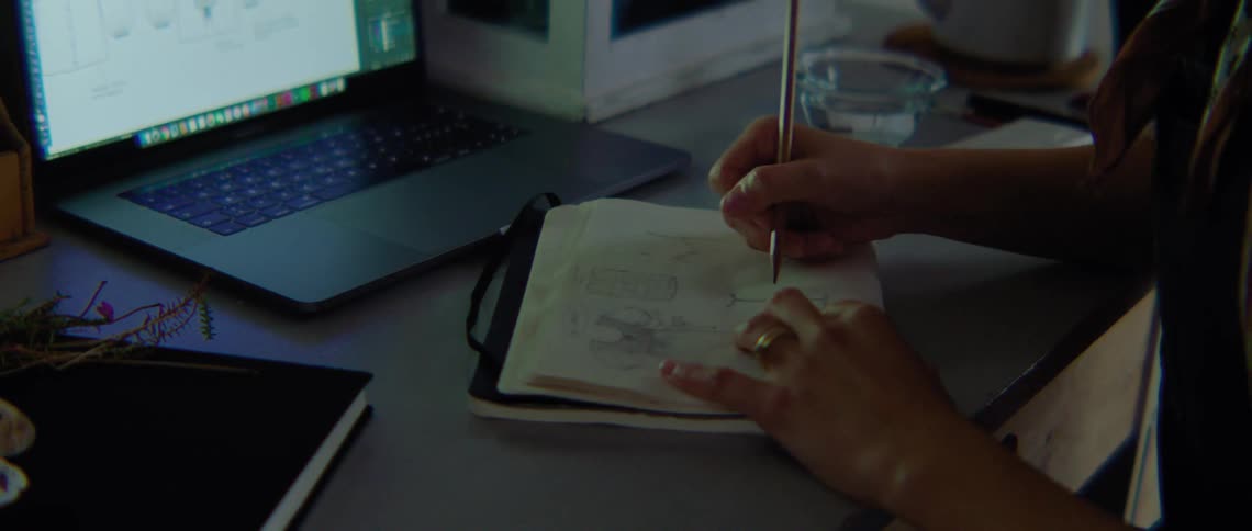 A woman is sitting at a desk and drawing on paper with a laptop opened during the day