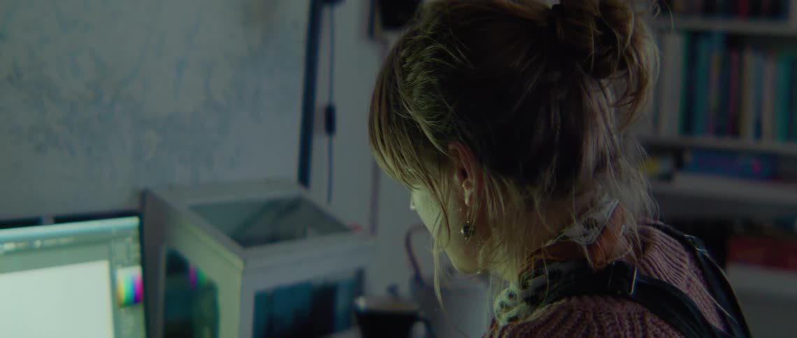 Woman with brown hair pink sweater and earrings sits at desk working on computer next to bookcase