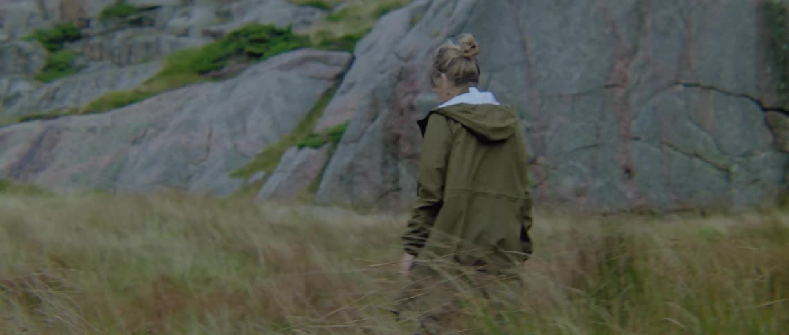 A woman walking in a grassy field with a cliff and trees in the background