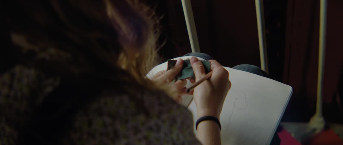 A woman is drawing in a book on her lap and handling pieces of fabric