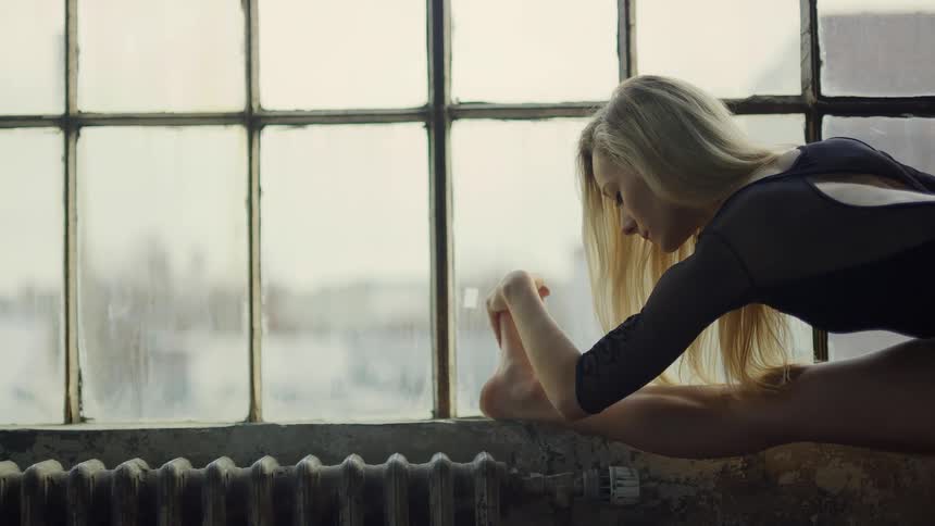 Blonde female ballerina with straight hair warms up before performing ballet near the window
