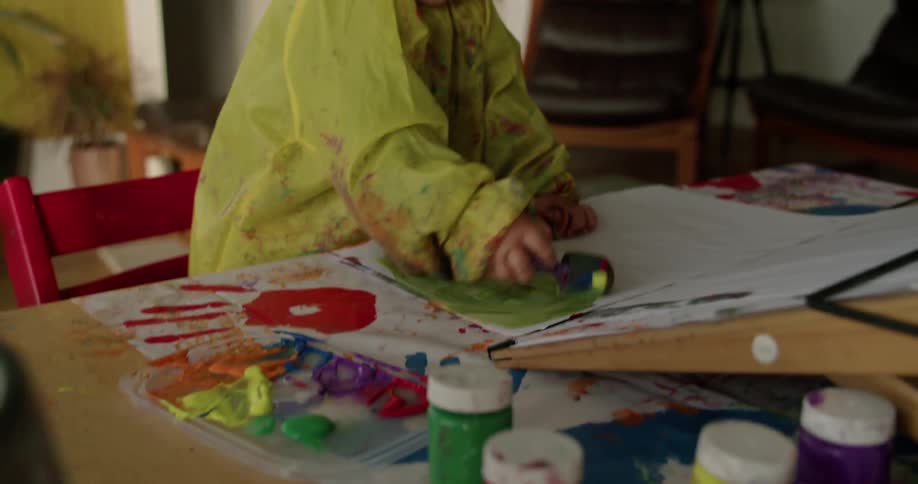 A little girl is painting on paper using a brush while surrounded with paints and supplies at home in the living room