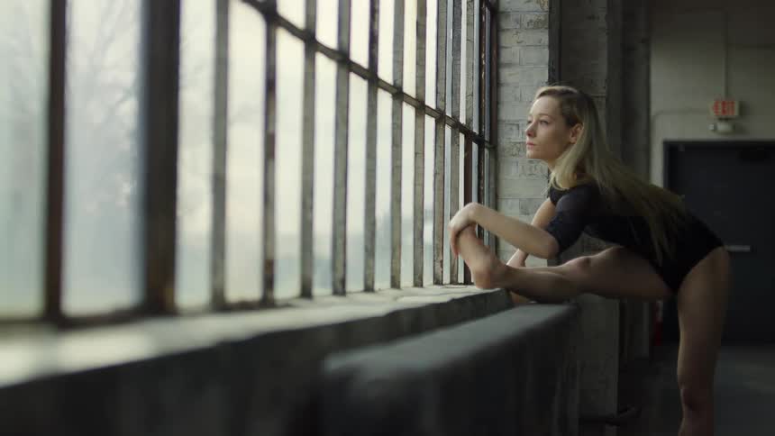 A dancer in a dance studio is stretching her legs on a window sill during the daytime