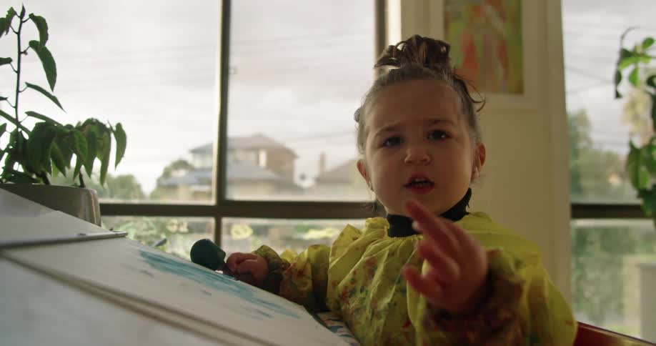 A child is sitting at a desk with painted paper and holding a sponge paint brush in one hand and holding up the other hand
