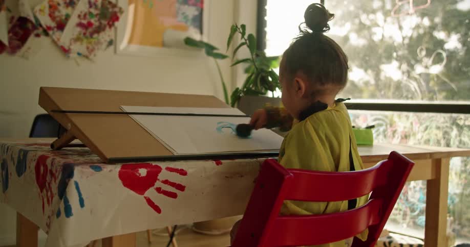 A child is sitting in a red chair and painting on paper with a sponge brush