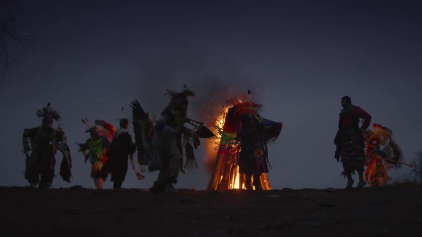 Native americans in traditional dress dance around a fire on a ridge