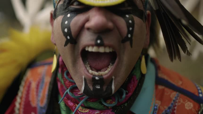 A native american man wearing a headdress and tribal costume turns around and yells