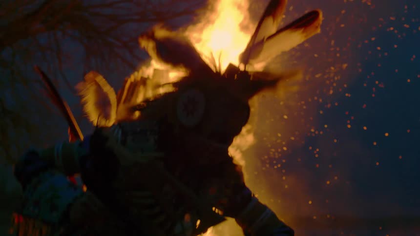 A large bonfire rages behind an indigenous dancer
