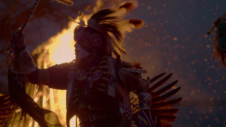 Native american people are wearing tribal clothing and dancing by a bonfire