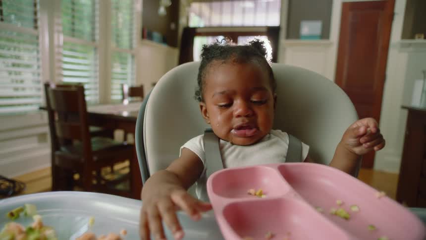 A baby sitting in a high chair in a kitchen is spilling a plate of food