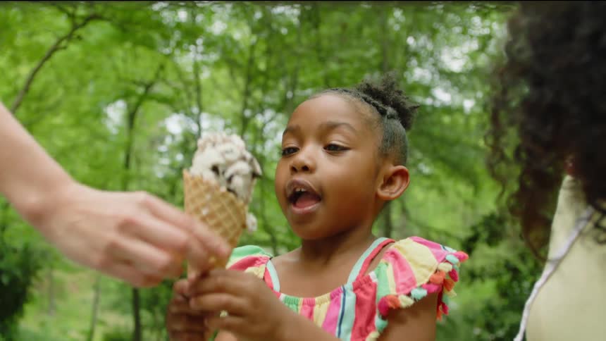 A woman brings an ice cream to a little girl who tries to eat it before the ice cream falls off the cone