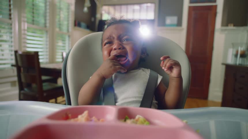 A baby sits in a highchair with food in front of her and cries