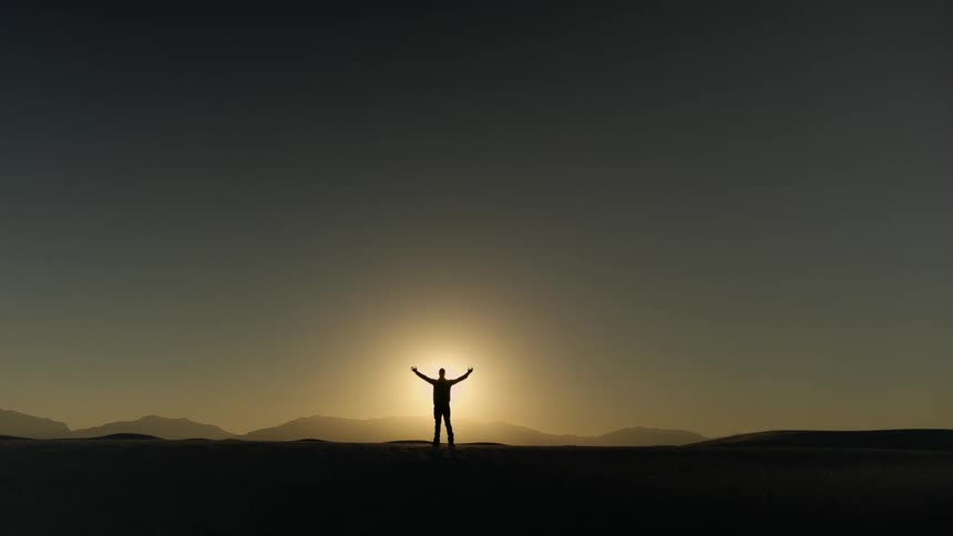 A silhouette of a person standing on a landscape with the sun the sky and clouds in the background