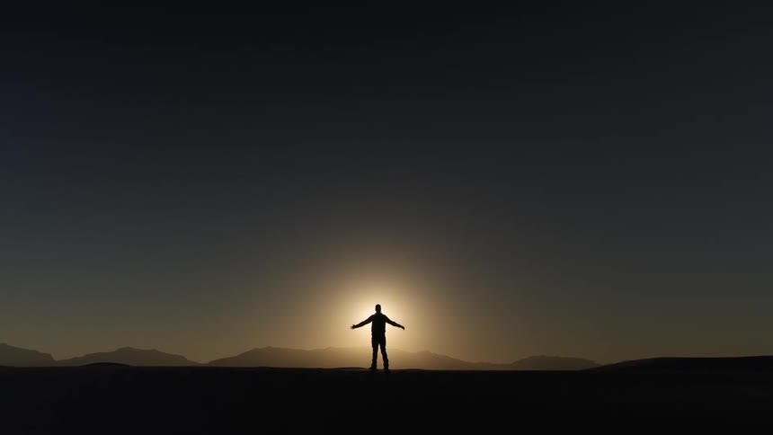 Silhouette of a man holding up his arms in front of the sun near the mountains