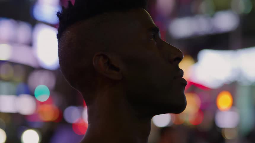 A young man stands outside and looks up at the bright city lights as cars drive past