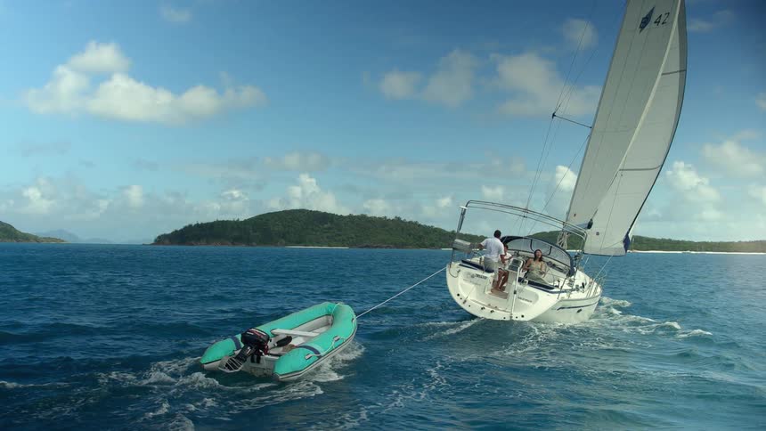 Two people are on a sailboat that is towing another water vessel