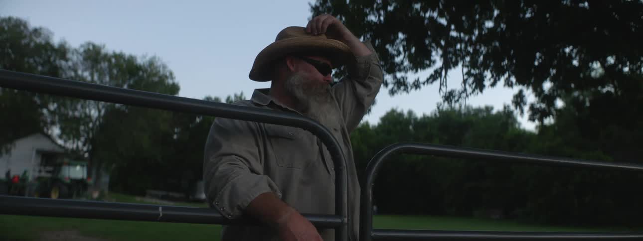 A cowboy stands with his arms on a gate reaching up to adjust his hat forward