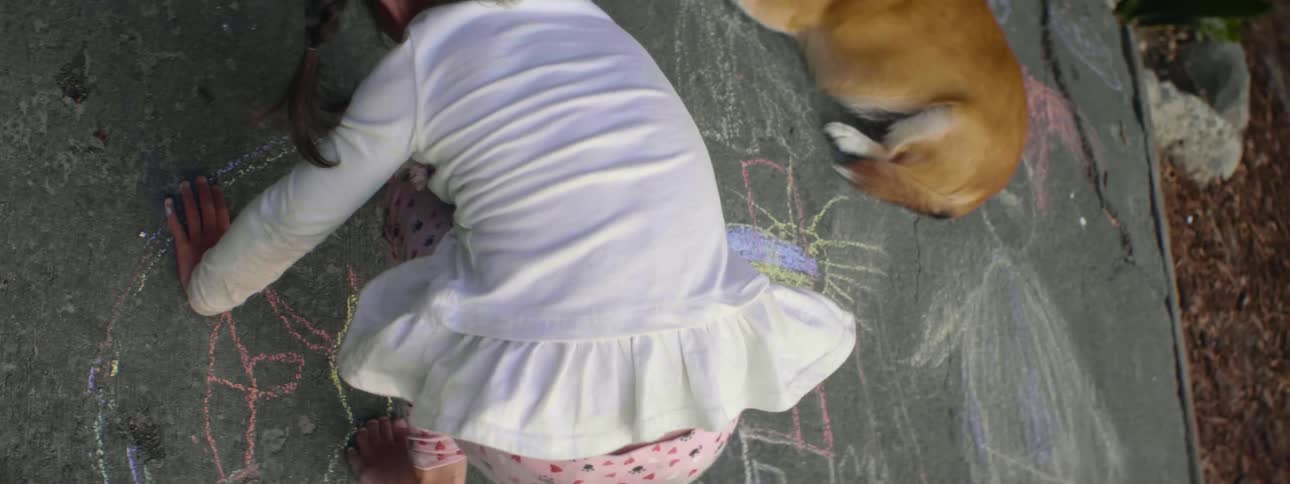 A child draws with chalk on the concrete ground as a dog lays down