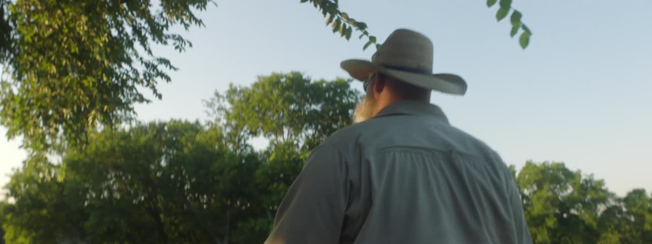 A man with a hat and sunglasses walking towards a barn with trees the sky and clouds in the background