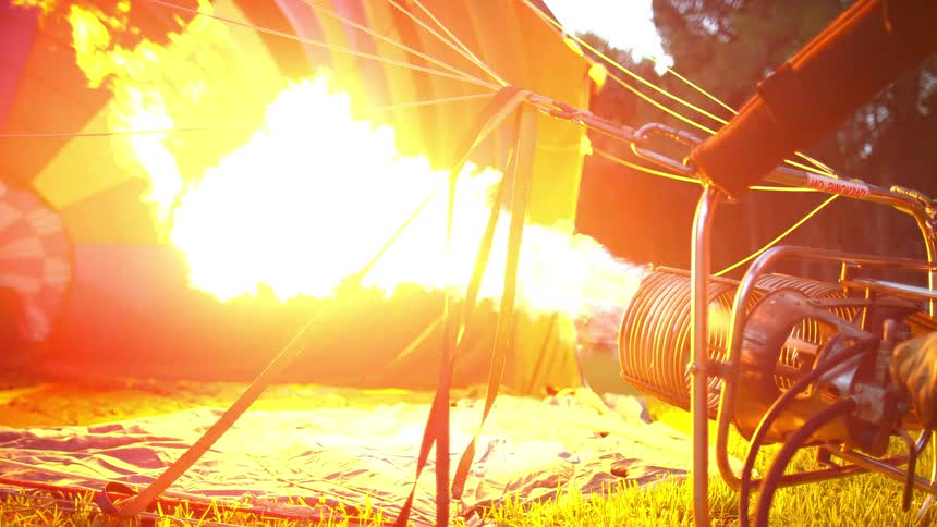 Person turns on flame heating air entering hot air balloon laying on grass near trees