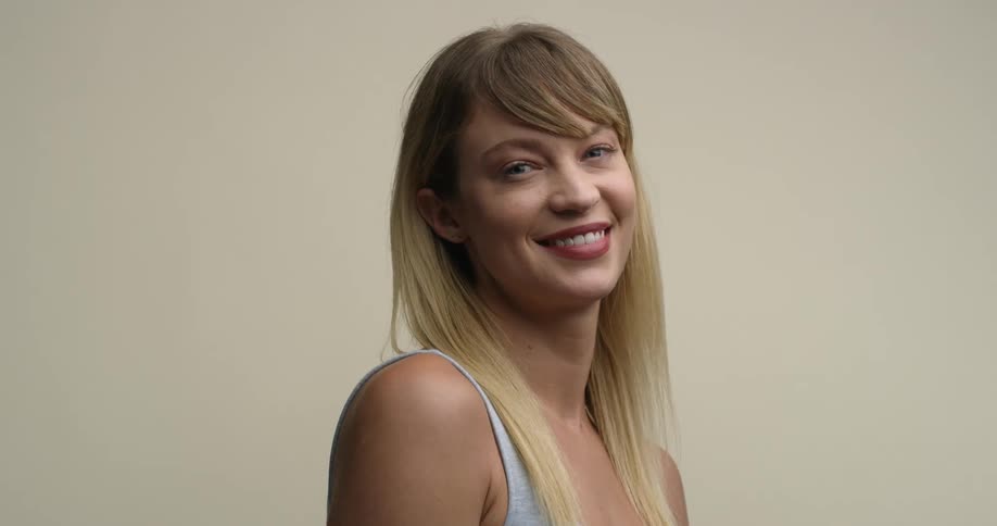 A young, blonde white woman with bangs/fringe turns from looking to the side to giving a direct smile to the camera, followed by a slight head tilt.