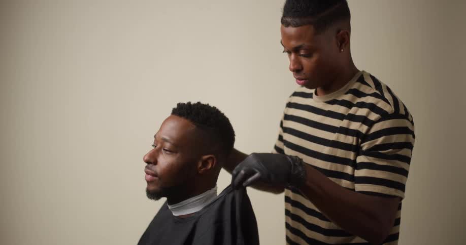 A black male barber putting a cape on a seated black male customer to prepare for a hair cut