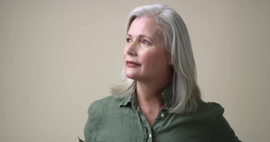 A beautiful aged white woman with dark eyes, red lips, and gray hair grasps her collar as she looks up and to the side, and then straight at the camera.
