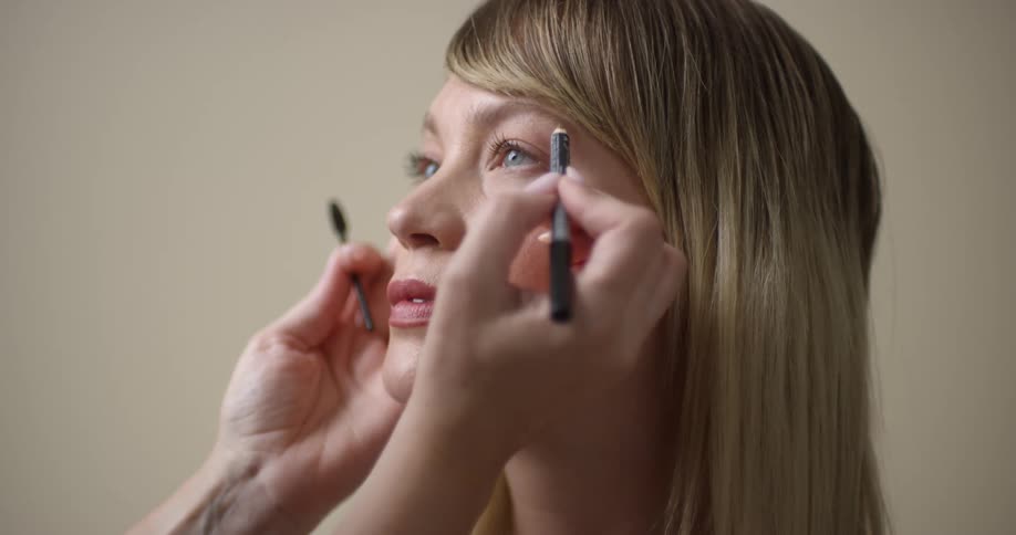 A manicured stylist aplying makeup on a young, blonde, Caucasian woman's eyebrows.