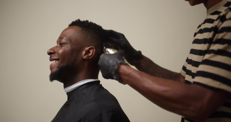 A young black man with a beard is getting a haircut from a young black male barber with an electric razor