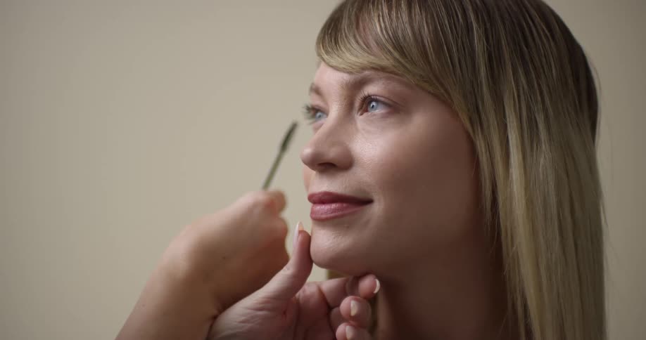 A young, blonde, white woman with light eyes is having her makeup done by a manicured esthetician.