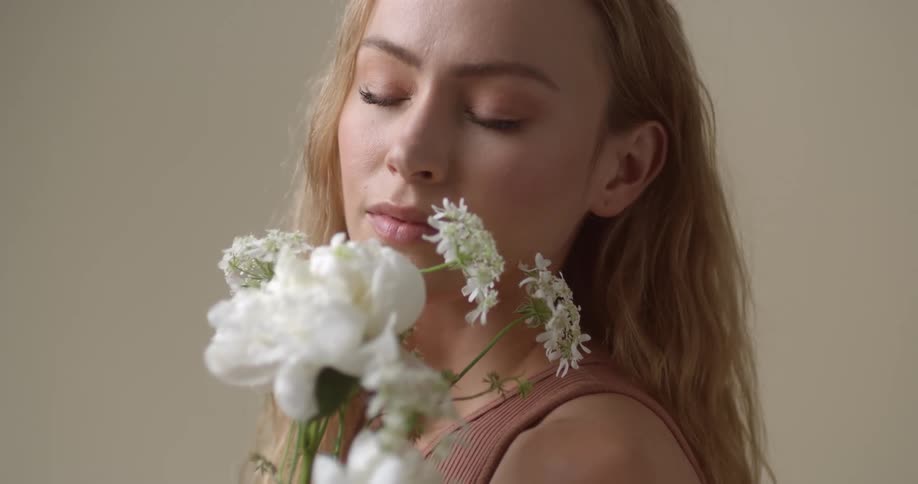 A young, blonde, Caucasian woman is smelling and enjoying the scent of a bouquet of white flowers with her eyes closed.