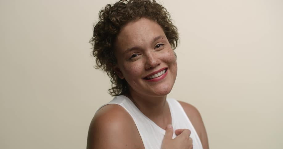 A young white woman with short hair and freckles posing with her hand over her heart and smiling