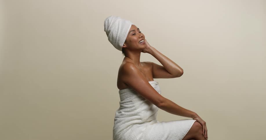 A young, multicultural woman is sitting down wearing towels on her head and body as she smiles and strokes her skin.