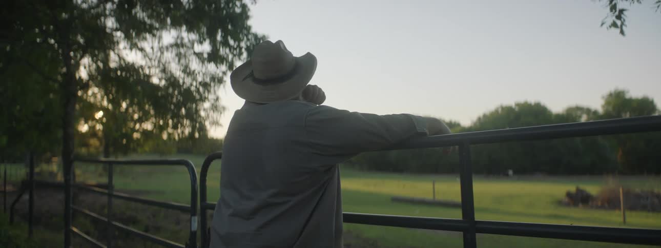 A heavy set man wearing sunglasses and a cowboy hat is leaning over a rail relaxing