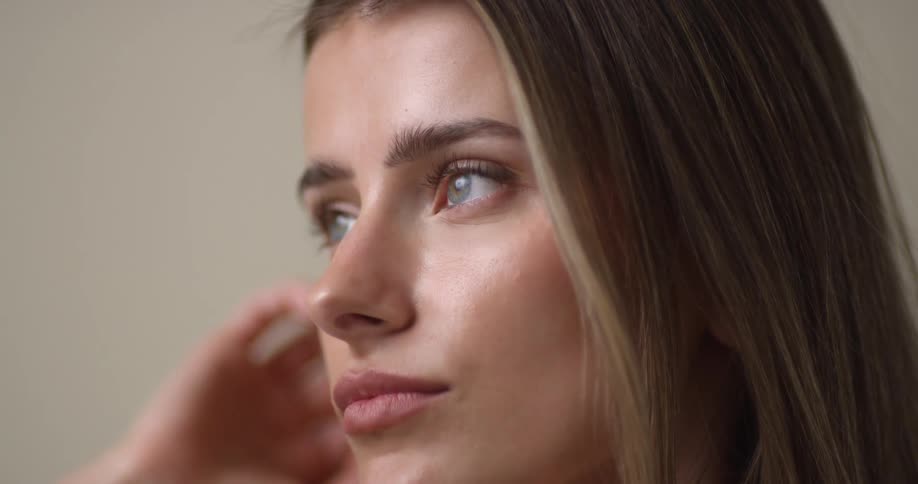 A young, Caucasian woman with light eyes pulls her dark-blonde hair back with one hand and glances to the other side with an aloof look.