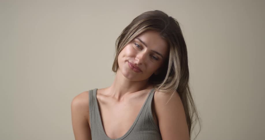 A young, white female poses seductively as she tosses her hair to the side with parted lips, and lifts one shoulder and then the other while giving an enticing smile.