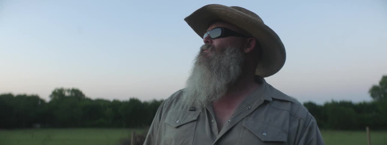 Man with cowboy hat sunglasses long beard and gray shirt looks around in the field at dusk