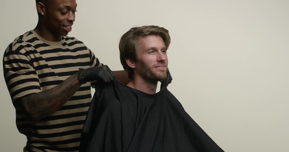 A young, black barber puts a cape around a young, blonde, white man sitting in a chair preparing for a haircut.