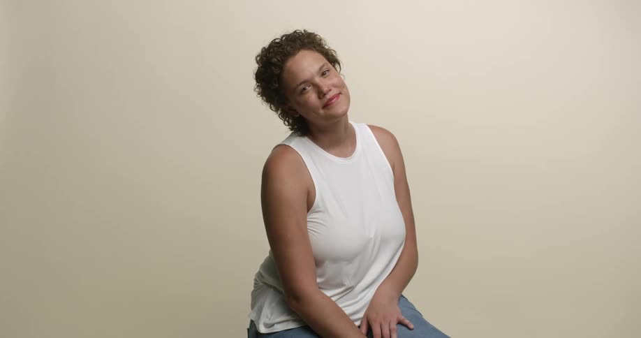 A young, freckled woman with short curly hair is sitting on a chair looking at the camera with her head tilted to one side and a slight smile