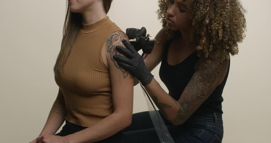 A caucasian woman is getting inked by a woman-of-color with a tattoo gun and big, beautiful, curly hair