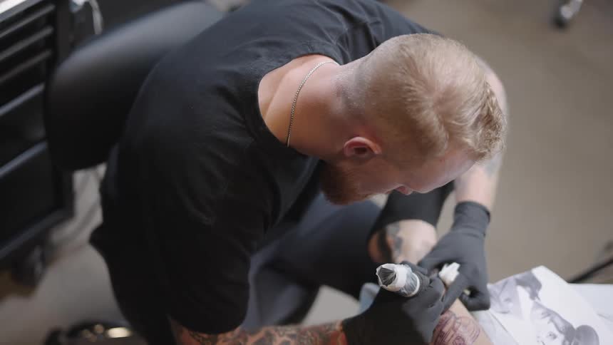 Blonde tattoo artist with black t shirt and gloves applies tattoo on mans arm by looking at photograph on tattoo studio