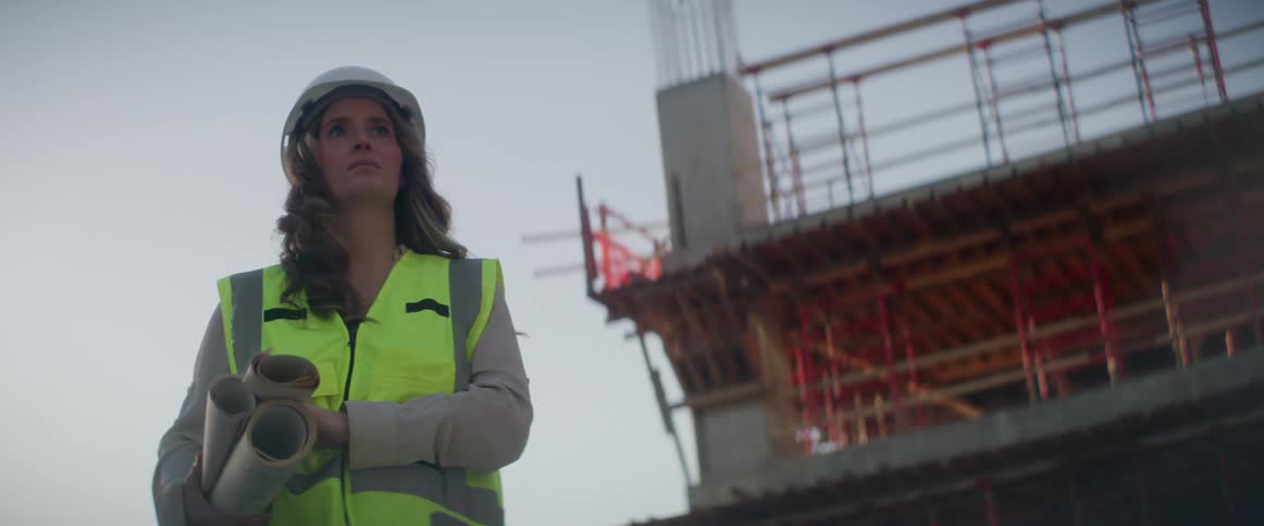 A woman carrying blueprints walks up at a construction site