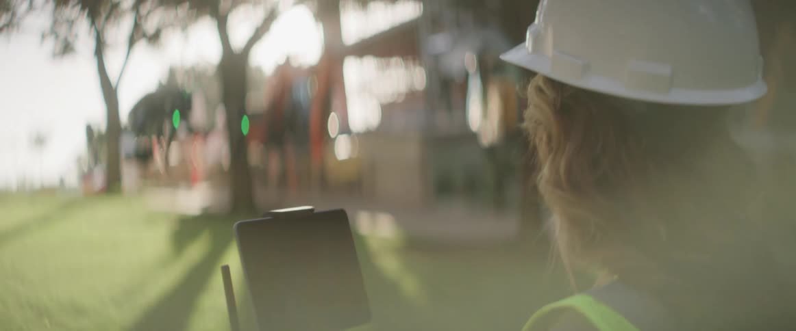 Female construction worker pilots drone through takeoff on lawn near building under construction