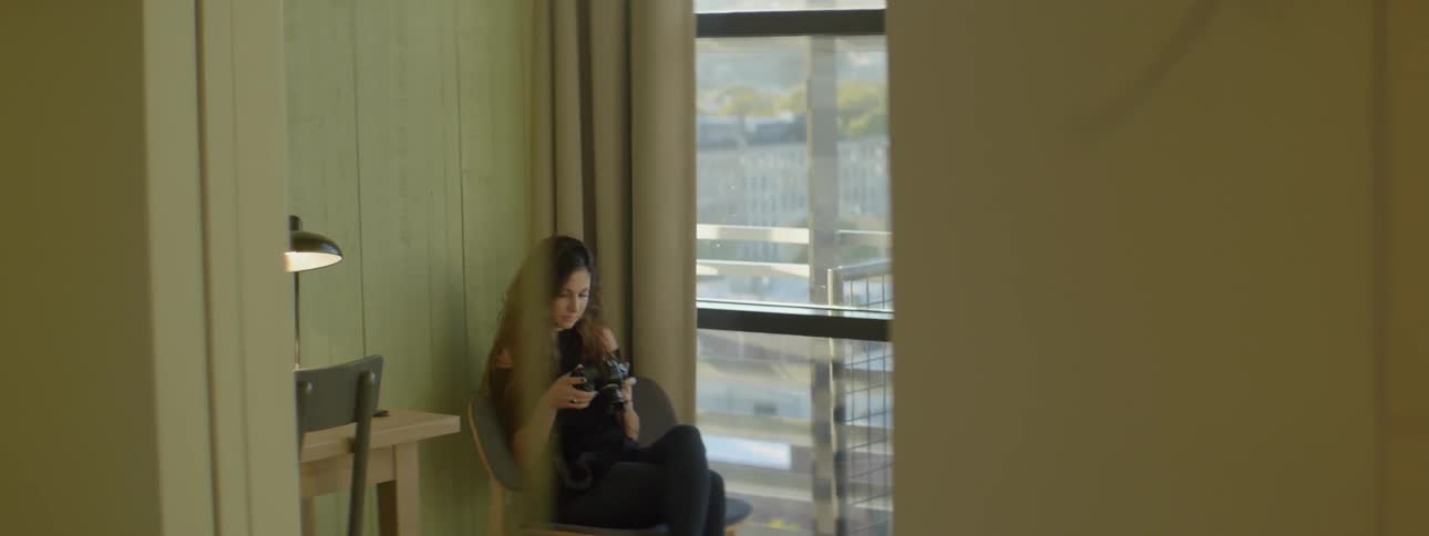 A woman is sitting near a window using a camera