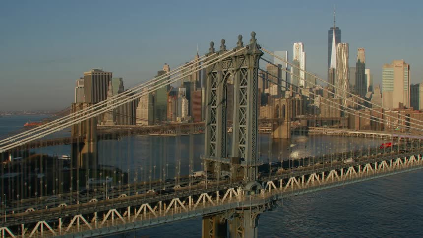 Cars travel across a bridge over a river to a big city with tall skyscrapers high in a clear blue sky
