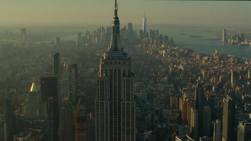 The spire on top of a skyscraper stands high above a big city with the skyline in the distance under a hazy sky